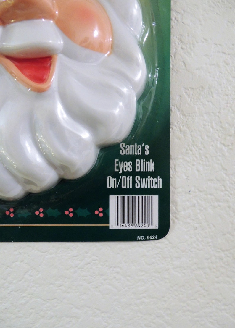 May include: A white plastic Santa Claus face with a red mouth and a white beard. The eyes blink on and off with a switch. The packaging is green with a barcode and the text "Santa's Eyes Blink On/Off Switch" and "NO. 6924".