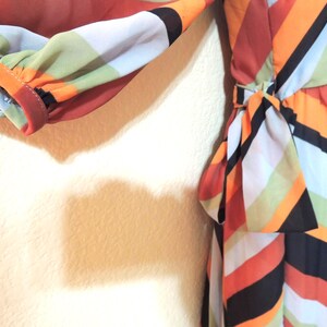 May include: A colorful dress with a bold geometric pattern. The dress features a mix of orange, black, white, and green stripes. The sleeve is gathered at the wrist. A bow detail is at the waist.
