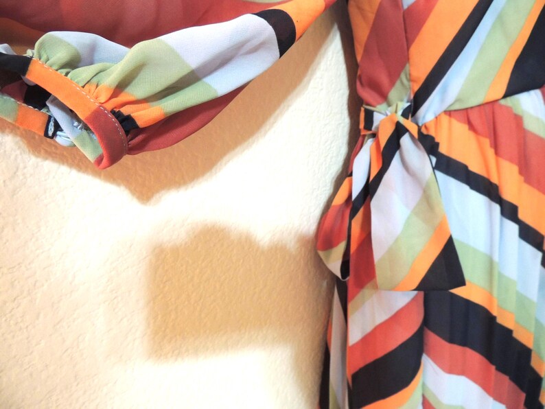 May include: A colorful dress with a bold geometric pattern. The dress features a mix of orange, black, white, and green stripes. The sleeve is gathered at the wrist. A bow detail is at the waist.