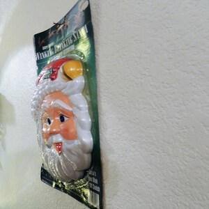 May include: A plastic Santa Claus head with a white beard and a red hat. The head is winking and has a light-up nose. The packaging says "Winking Blinking Santa".