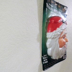 May include: A plastic, white and red, winking Santa Claus head with a white beard and a red hat. The packaging says "Winking Blinking Santa" and "Merry Christmas".