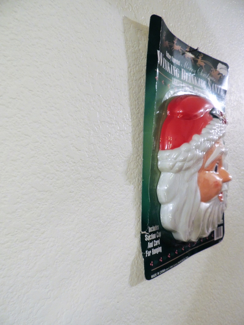 May include: A plastic, white and red, winking Santa Claus head with a white beard and a red hat. The packaging says "Winking Blinking Santa" and "Merry Christmas".