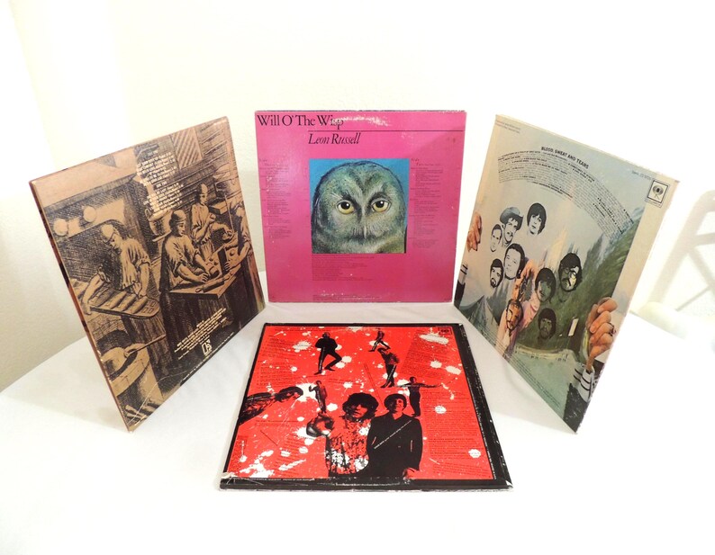 May include: Four vintage vinyl record album covers. One pink album cover features an owl's face, and the text "Will O' The Wisp" and "Leon Russell". Another album cover is red with black silhouettes. The other two album covers have illustrations.