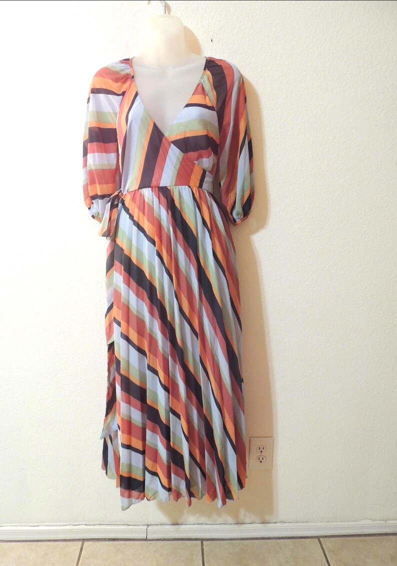 May include: A long, pleated dress with a V-neck and long sleeves. The dress features vertical stripes in shades of orange, brown, black, and blue. The dress is displayed on a mannequin.