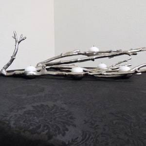 Puede incluir: Portavelas de metal plateado con forma de rama de &aacute;rbol. El dise&ntilde;o de la rama sostiene cinco velas de t&eacute; blancas. El portavelas se exhibe sobre una superficie negra.