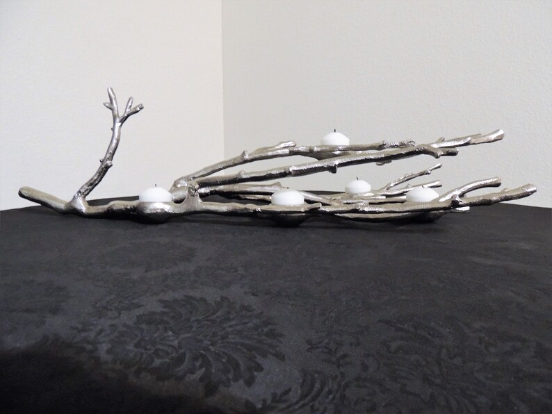 Puede incluir: Portavelas de metal plateado con forma de rama de &aacute;rbol. El dise&ntilde;o de la rama sostiene cinco velas de t&eacute; blancas. El portavelas se exhibe sobre una superficie negra.