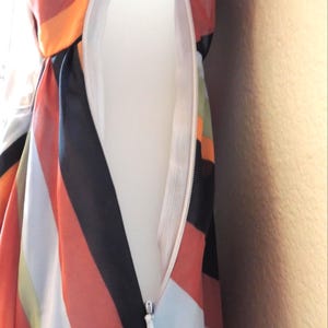 May include: Close-up of a colorful dress with a white zipper. The dress features bold stripes in orange, black, white, and green. The zipper is white and partially open, revealing the inside lining of the dress.