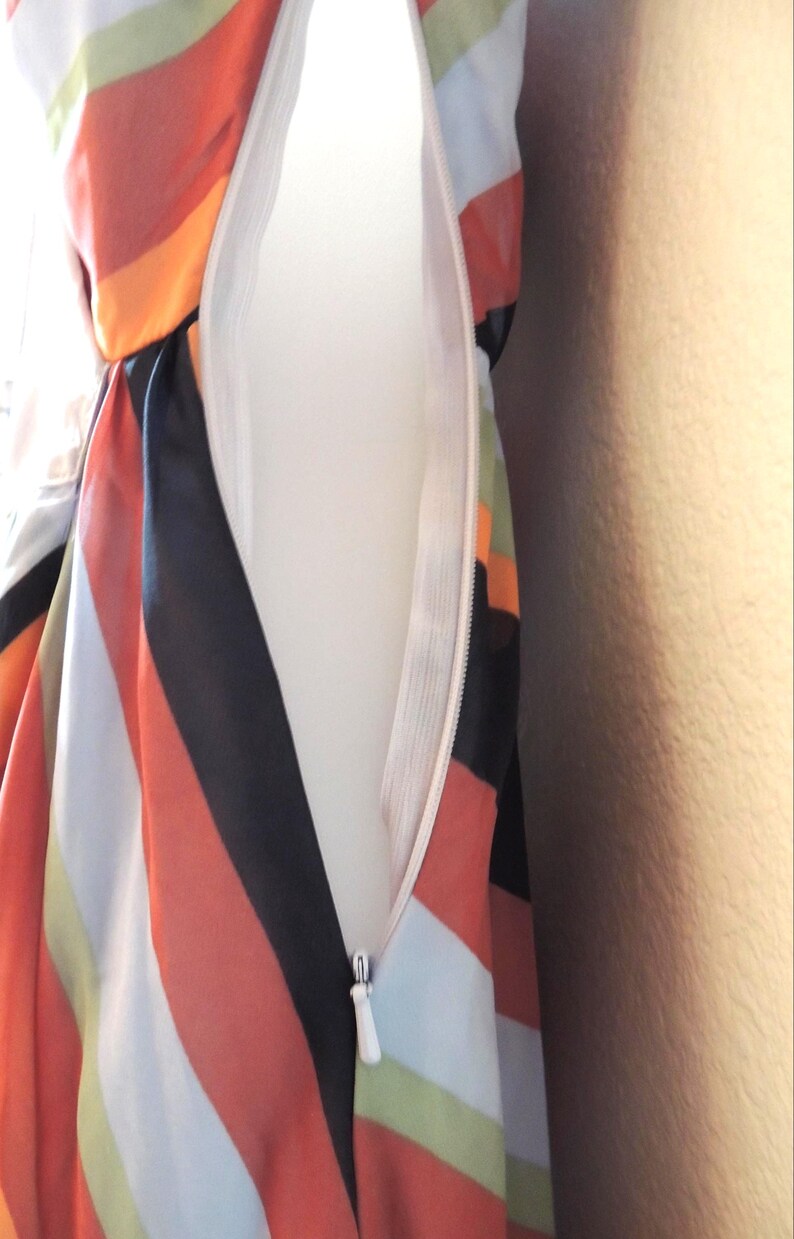 May include: Close-up of a colorful dress with a white zipper. The dress features bold stripes in orange, black, white, and green. The zipper is white and partially open, revealing the inside lining of the dress.