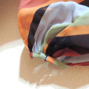 May include: Close-up of a colorful, striped fabric. The fabric features horizontal bands of orange, black, white, green, and peach. The sleeve is gathered at the cuff with a small button detail. The material appears to be lightweight and sheer.