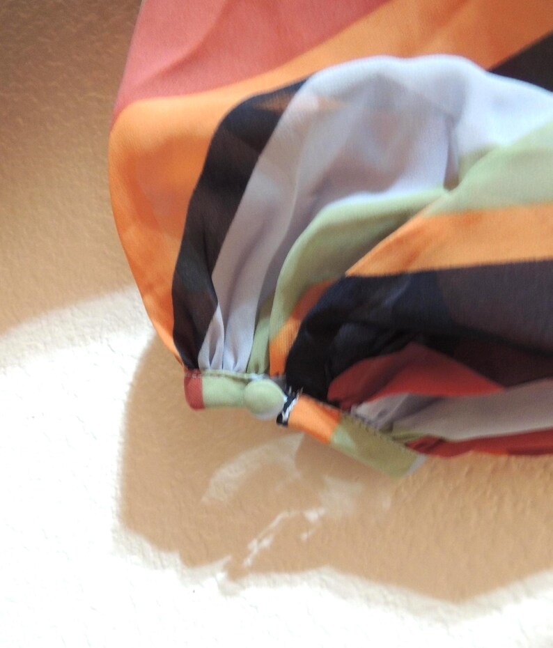 May include: Close-up of a colorful, striped fabric. The fabric features horizontal bands of orange, black, white, green, and peach. The sleeve is gathered at the cuff with a small button detail. The material appears to be lightweight and sheer.
