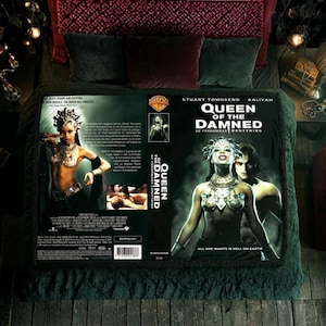 Puede incluir: Una manta decorativa con el cartel de la película "Queen of the Damned", con Aaliyah y Stuart Townsend. La manta se muestra en una cama con ropa de cama verde oscuro y burdeos. El título de la película es visible.