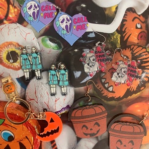 May include: A collection of Halloween-themed earrings featuring characters from horror movies. The earrings are made of wood and acrylic and feature various designs, including a ghost, a pumpkin, and a scream mask. The earrings are all in different colors, including black, orange, white, and purple. The earrings are displayed on a background of candy and other Halloween decorations.