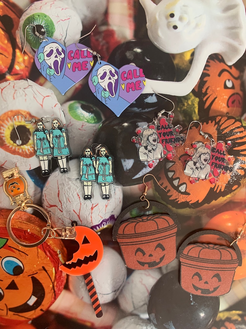 May include: A collection of Halloween-themed earrings featuring characters from horror movies. The earrings are made of wood and acrylic and feature various designs, including a ghost, a pumpkin, and a scream mask. The earrings are all in different colors, including black, orange, white, and purple. The earrings are displayed on a background of candy and other Halloween decorations.