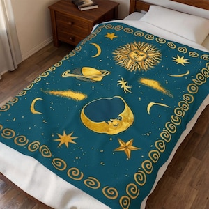 May include: A teal blanket with a celestial design featuring a smiling moon, sun, stars, planets, and comets in gold. The blanket has a gold border with a repeating swirl pattern. The blanket is on a bed with a white sheet.