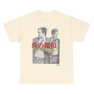 Pode incluir: Camiseta creme com uma imagem em preto e branco de dois homens de terno. O texto japonês "True Detective" é impresso em vermelho e preto acima da imagem. A camiseta tem gola redonda e mangas curtas.