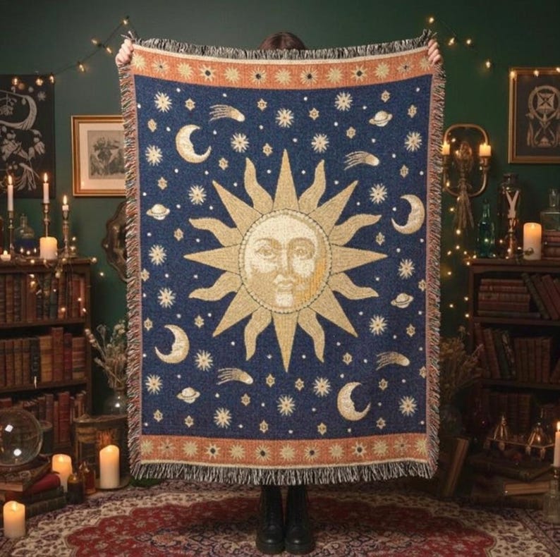 May include: A woven throw blanket featuring a large central sun face with golden rays surrounded by cream-colored moons, stars, and planets against a deep navy blue background, finished with a decorative orange border and fringed edges.
