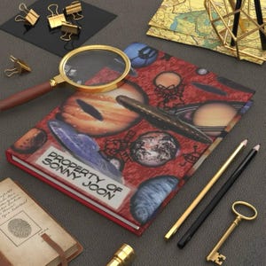 Puede incluir: Un diario rojo con un diseño temático espacial con planetas y el texto "PROPERTY OF SONNY JOON". También hay una lupa con borde dorado, lápices y una llave. La escena incluye clips dorados y un pequeño cuaderno.