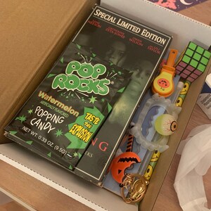May include: A box filled with Halloween themed items, including a pack of watermelon flavored Pop Rocks candy, a glow-in-the-dark eyeball, a plastic vampire teeth, a Rubik's cube, and a plastic jack-o-lantern keychain. The box also contains a DVD case for the movie "The Ring" starring Naomi Watts.