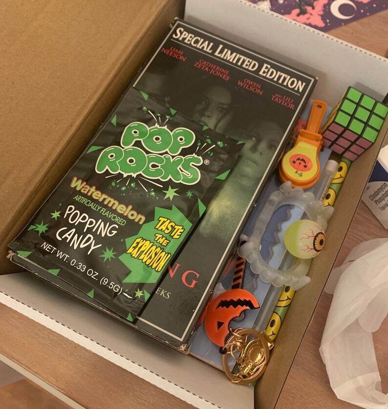 May include: A box filled with Halloween themed items, including a pack of watermelon flavored Pop Rocks candy, a glow-in-the-dark eyeball, a plastic vampire teeth, a Rubik's cube, and a plastic jack-o-lantern keychain. The box also contains a DVD case for the movie "The Ring" starring Naomi Watts.
