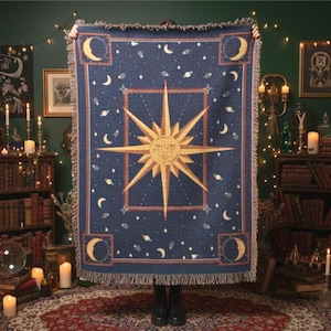 Peut inclure: Une couverture bleu foncé avec un motif céleste. La couverture présente un grand éclat de soleil au centre, entouré de lunes, d'étoiles et de planètes. La bordure a un cadre rouge et des franges.