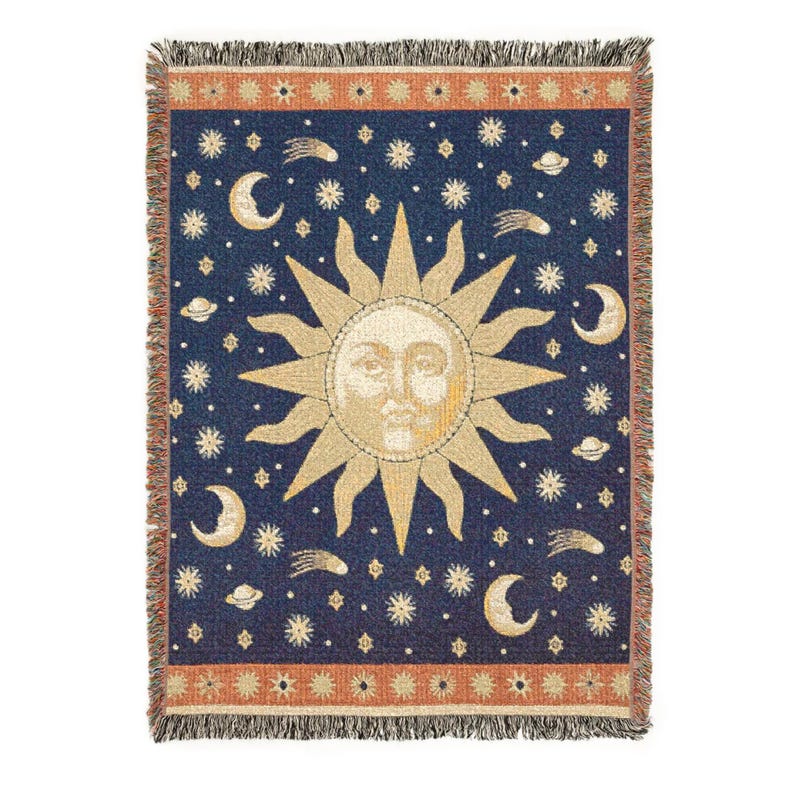 May include: A woven throw blanket featuring a central sun face with rays on a dark blue background. The design includes stars, crescent moons, and planets scattered throughout. The top and bottom borders are orange with a repeating star pattern.