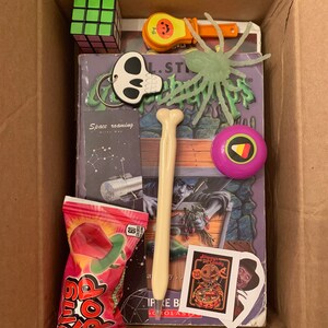 May include: A cardboard box filled with Halloween themed items, including a book, a green spider toy, a candy, a bone-shaped pen, a small pumpkin, a Rubik's cube, a skull keychain, and a purple and yellow ball.