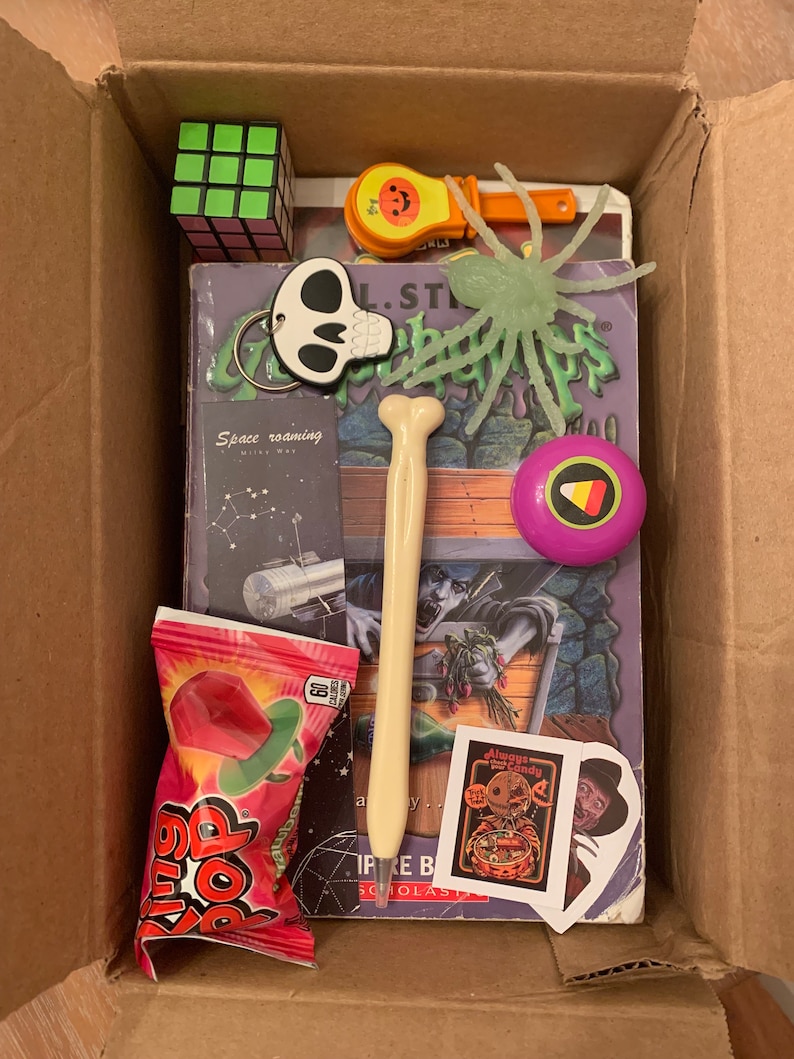 May include: A cardboard box filled with Halloween themed items, including a book, a green spider toy, a candy, a bone-shaped pen, a small pumpkin, a Rubik's cube, a skull keychain, and a purple and yellow ball.