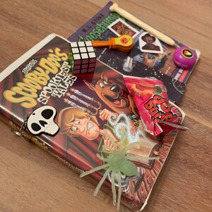 May include: A Scooby-Doo VHS tape with a spooky theme, a green plastic spider, a Rubik's cube, a candy wrapper, a book titled "Goosebumps", a bone-shaped pencil, a small plastic pumpkin, and a purple and yellow sticker.