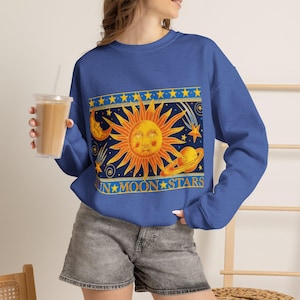 May include: A royal blue long-sleeve sweatshirt featuring a large rectangular graphic print of a yellow sun with a face, a crescent moon, a planet with rings, and stars. The text SUN MOON STARS is printed in yellow at the bottom of the graphic.