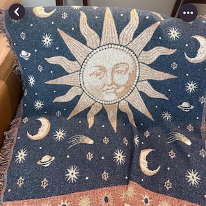 May include: A woven throw blanket featuring a dark blue background with a large central sun face in beige and gold. The design includes scattered stars, crescent moons, planets, and comets. A decorative border with sun motifs runs along the bottom edge.