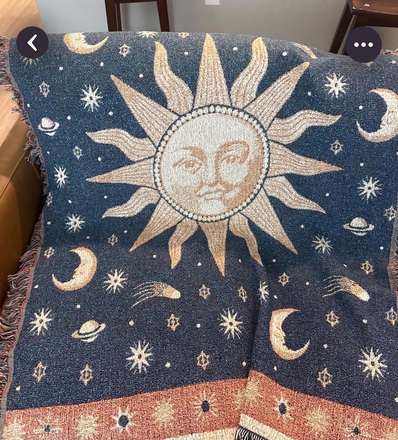 May include: A woven throw blanket featuring a dark blue background with a large central sun face in beige and gold. The design includes scattered stars, crescent moons, planets, and comets. A decorative border with sun motifs runs along the bottom edge.