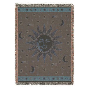 May include: A rectangular woven throw blanket featuring a central sun face with radiating rays, surrounded by crescent moons, stars, and comets. The design uses a brown background with blue and cream accents, finished with a fringed edge on all sides.