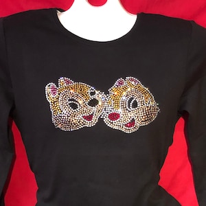 May include: Black long-sleeve shirt featuring a rhinestone design of two cartoon chipmunks. The chipmunks have brown, gold, red, and white rhinestones. The shirt is displayed on a white hanger against a red background.