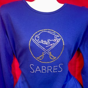 May include: A royal blue long-sleeve shirt featuring the Buffalo Sabres logo in gold and white rhinestones. The word "Sabres" is spelled out in white rhinestones below the logo. The shirt is displayed on a mannequin.