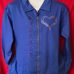 May include: Royal blue zip-up jacket with a white zipper and a heart design. The word "Buffalo" is spelled vertically in red and white rhinestones. The heart design is also made of red and white rhinestones.