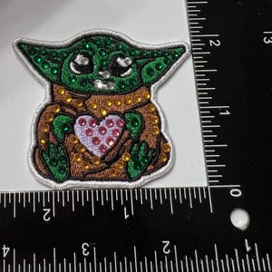 May include: Embroidered patch featuring a green alien-like character with large eyes and a heart-shaped design on its chest. The patch is embellished with various colored rhinestones and has a white border.