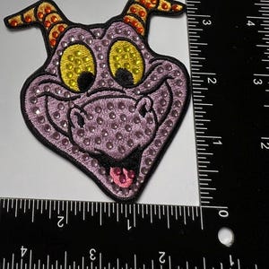 May include: Embroidered patch featuring a purple dragon head with yellow eyes and orange horns. The dragon's face is covered in small, sparkling rhinestones. The patch is approximately 3 inches wide.