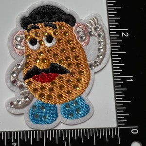 May include: Embroidered patch featuring a cartoon character with a tan face, black hair, a black mustache, and a red mouth. The character's face is covered in gold rhinestones, with silver rhinestones around the ears. The character is wearing blue shoes.