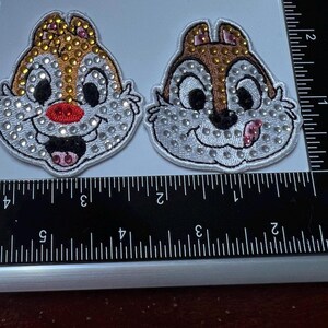 May include: Two embroidered patches featuring cartoon chipmunk faces. One has a red nose and open mouth, the other has a pink tongue sticking out. Both have brown and gold accents and are embellished with rhinestones.