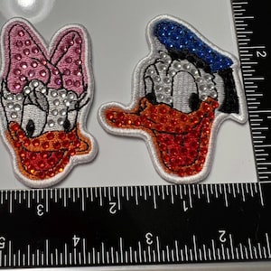 May include: Two embroidered patches featuring cartoon characters. One patch depicts a character with a pink bow, and the other features a character with a blue hat. Both patches are embellished with rhinestones and have red, white, and black accents.