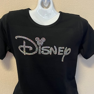 May include: Black t-shirt with the word "Disney" spelled out in iridescent rhinestones. The "i" in Disney is replaced with a Mickey Mouse head. The shirt has short sleeves and a crew neck.