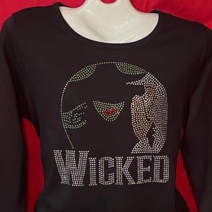 May include: Black long-sleeved shirt with a green, red, and silver rhinestone design of a witch's face and the word "WICKED".