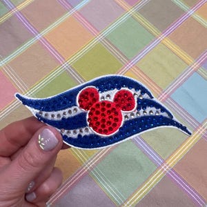 May include: A blue and white patch with a red Mickey Mouse head design. The patch is embellished with rhinestones and held by a hand with a floral nail design. The background is a colorful plaid pattern.