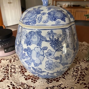 May include: A blue and white ceramic jar with a lid, featuring a floral pattern. The jar has a bulbous shape and a small handle on the lid. The floral design includes various flower types in shades of blue.