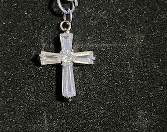 May include: A silver-colored necklace with a pendant in the shape of a cross. The cross is made of clear, rectangular-cut stones, with a round stone at the center. The chain is a linked design, and the background is black.