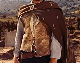 May include: A man wearing a brown cowboy hat, a brown suede vest, a brown poncho, and blue jeans. He is holding a gun in his right hand and has a cigar in his mouth.