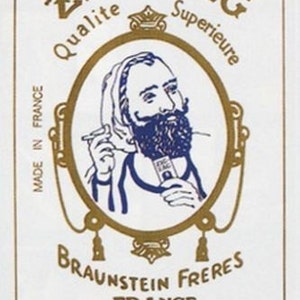 May include: A vintage Zig Zag cigarette paper advertisement featuring a man with a beard and a turban, holding a cigarette. The text reads "Zig Zag Qualite Superieure Braunstein Freres France Cigarette Papers".