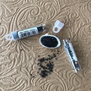 May include: Two small clear tubes filled with black seed beads. The tubes have labels with the text "680 DB00-0002V" and "Size 10/0". The beads are scattered on a brown textured surface.