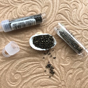 May include: Two small clear tubes filled with black seed beads. The tubes have labels with the text "690 DB00-0007V". The beads are scattered on a brown surface.