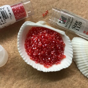May include: A display of small, red, cylindrical beads, labelled as "11/0" and "RD Light Siam", are presented in a white seashell. The beads are scattered, with some in clear plastic tubes. The image includes text labels and a white lid.